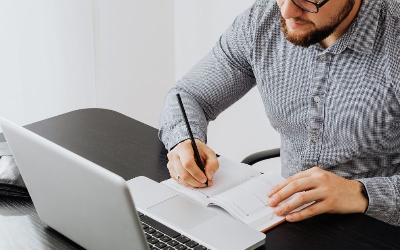 man in grey tshirt using a macbook pro and writing on a notebook